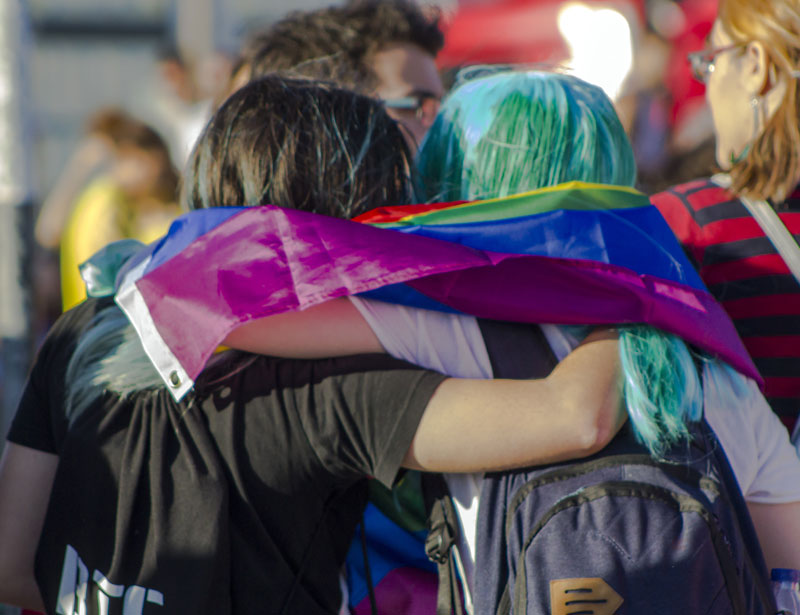 Zwei junge Personen haben sich gegenseitig einen Arm um die Schulter gelegt. Sie haben eine Regenbogenfahne umgehängt und gehen in einer Menschenmenge. 